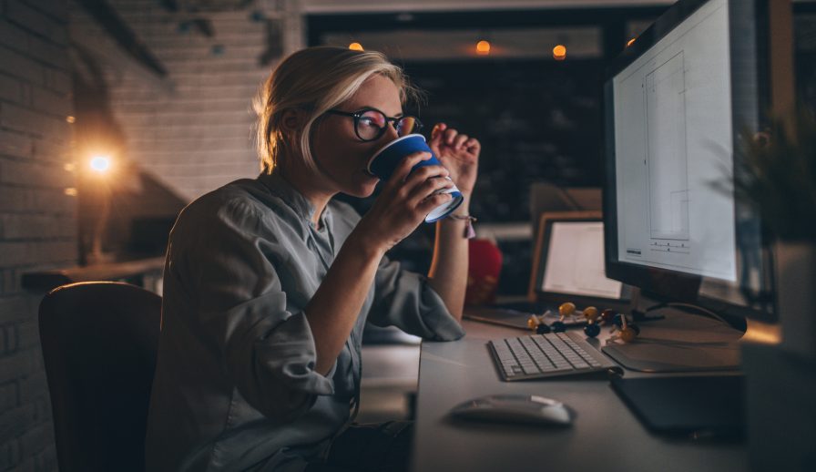 A woman sitting in front of a desktop computer screen drinking a coffee with a light on in the background